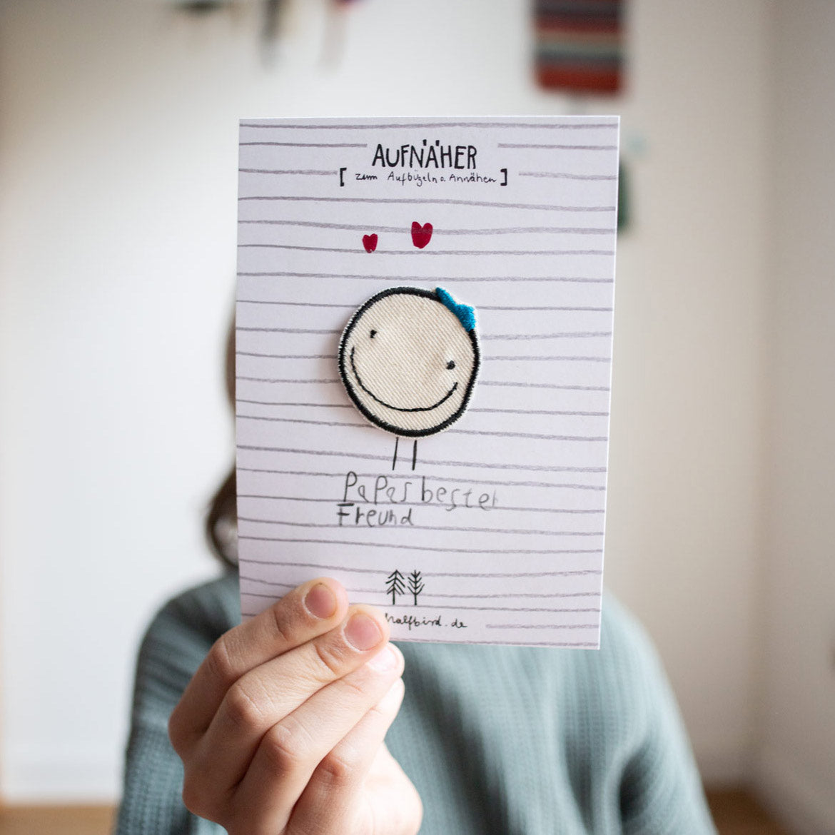 Kind hält eine Karte zum Muttertag und Vatertag hoch auf dem ein Smiley Aufnäher ist mit Beinen und einem kleinen Text darunter