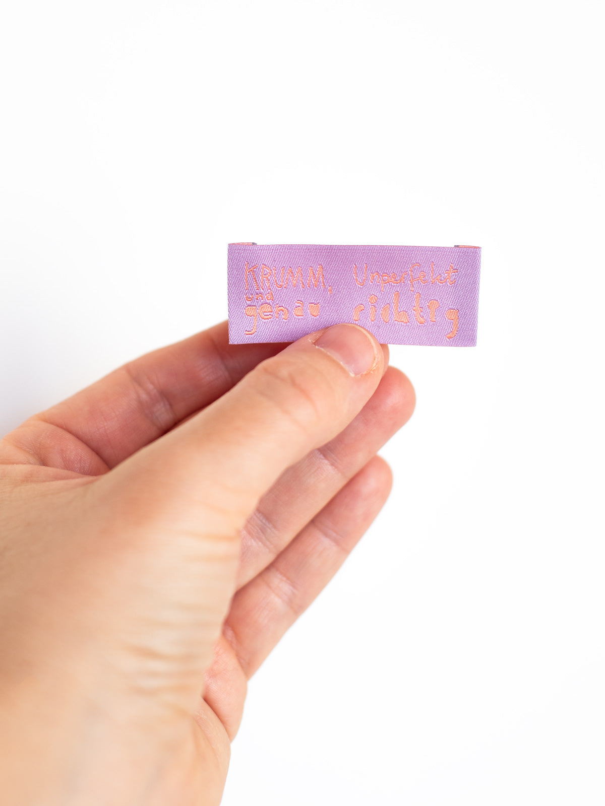 Hand mit fliederfarbenem Nähetikett mit der Aufschrift "krumm, unperfekt und genau richtig" von Halfbird