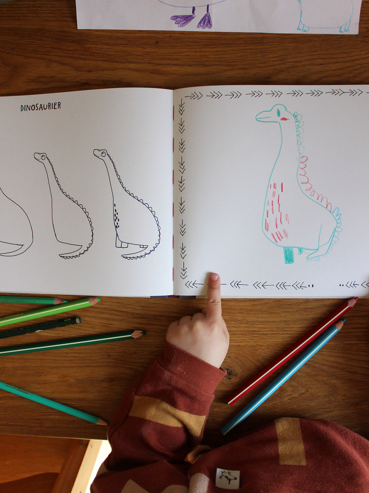 Kind zeigt auf selbstgemalten Dino in einem Buch. 