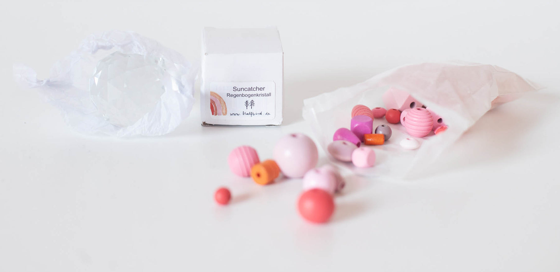 Auf dem tisch liegt eine papiertüte mit rosa perlen in verschiedenen Formen und nuancen, daneben liegt ein suncatcher kristall in rund von halfbird