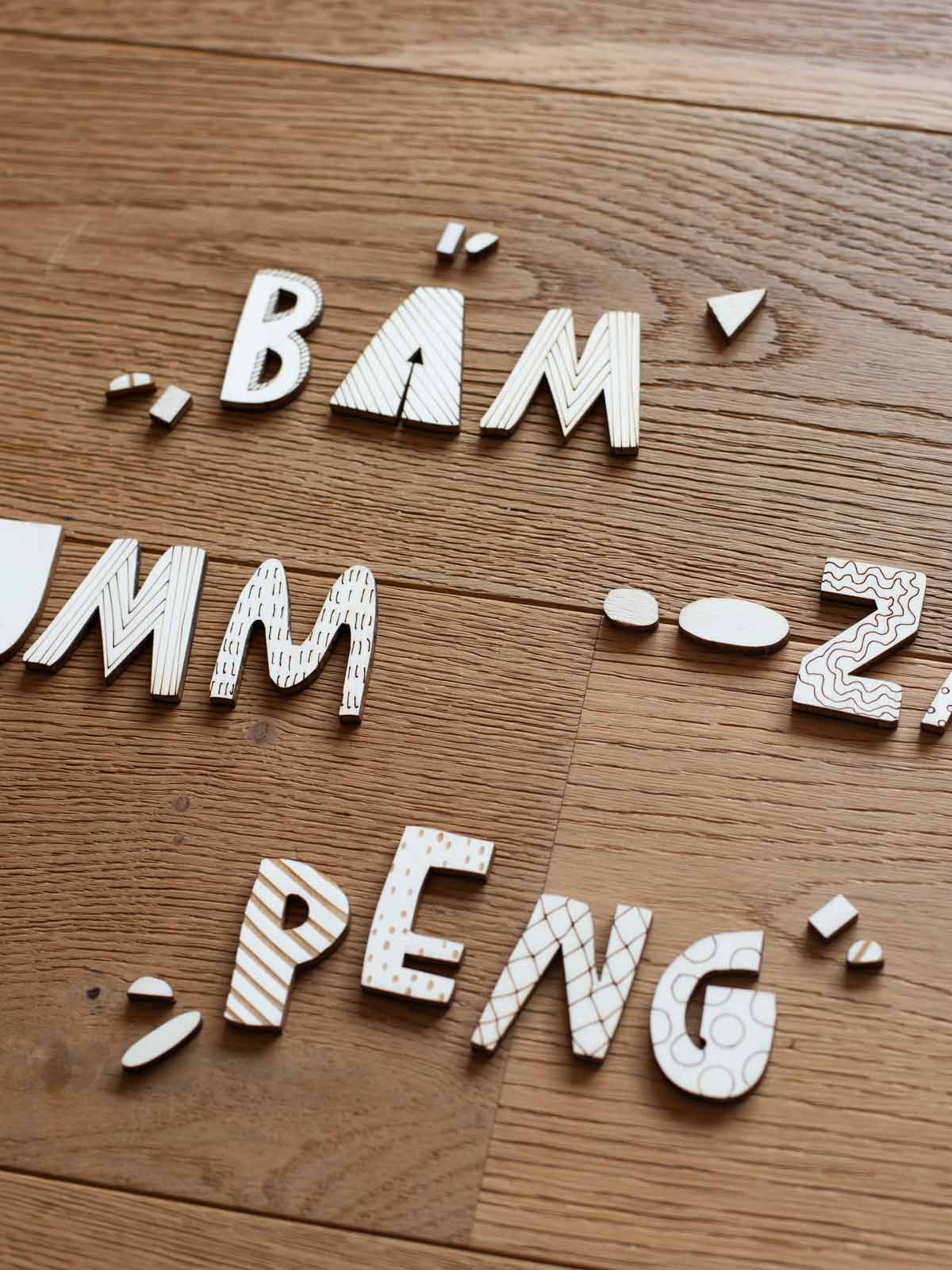 auf einem Holzboden liegen die Wörter Bäm, Bumm, Zack peng mit Holzbuchstaben in natürlicher Optik und verschiedenen eingelaserten Mustern