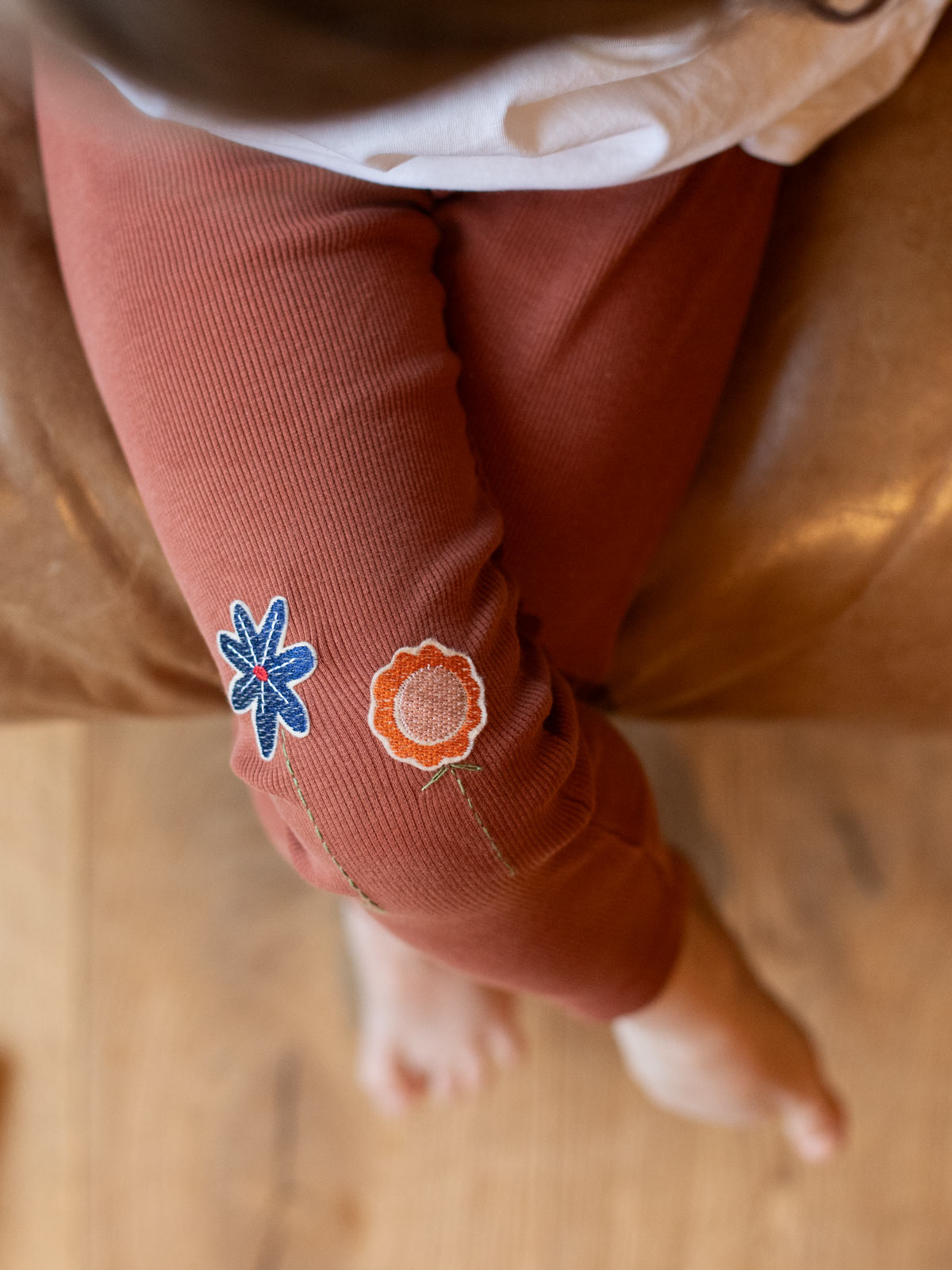 Kind trägt eine rostfarbene ripp leggings und darauf sind Blumen patches in blau und orange mit gestickten grünen Blütenstängeln