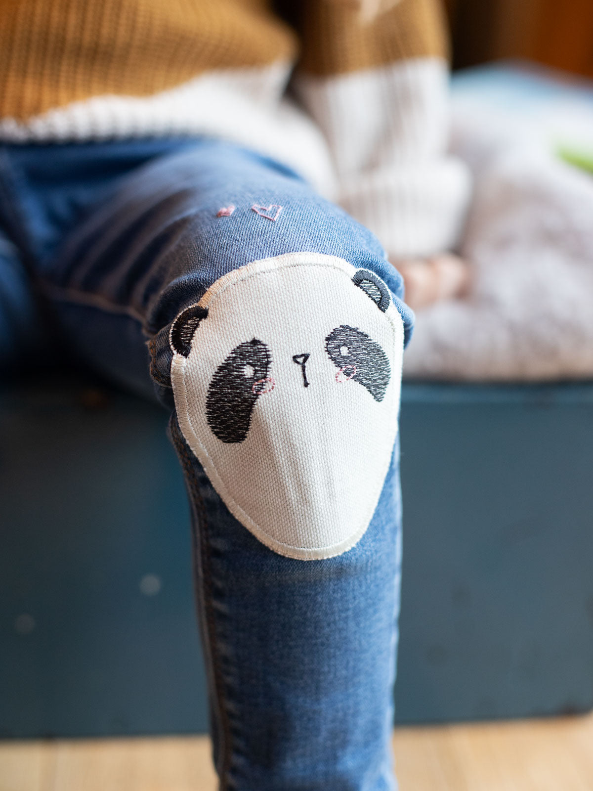 Kind sitzt auf dem Bett und trägt eine jeans, die mit einem Knieflicken als panda gesicht geflickt wurde