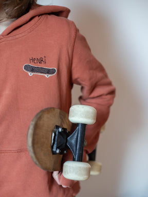 Kind trägt einen rostroten Hoodie mit einem gestickten Aufnäher Skateboard darauf und darüber ist sein Name gestickt. er hält ein Skateboard in der Hand