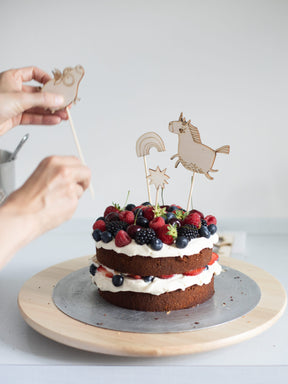Torte mit Beeren wird mit Cake Toppern aus Holz verziert. ein schwein, ein Einhorn, regenbogen und stern