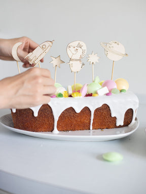 eine hand steckt einen weltraum caketopper auf einen verzierten Kuchen. 