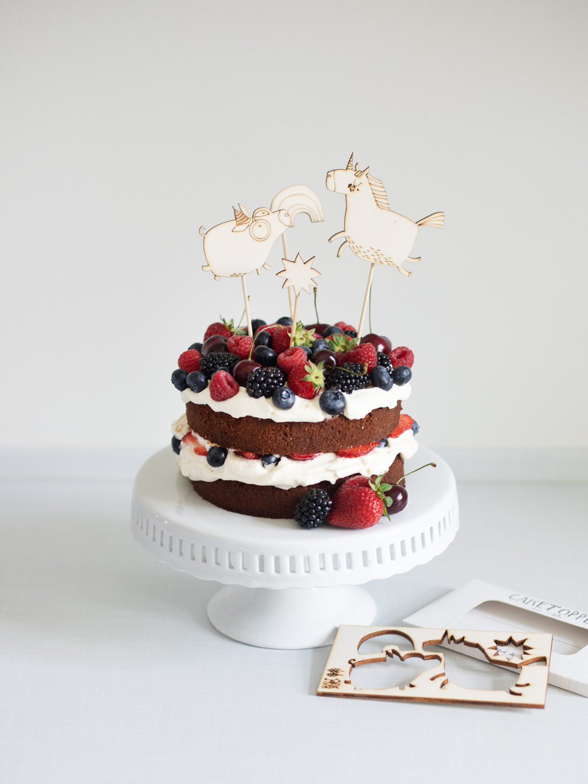 Geburtstagstorte verziert mit Erdbeeren, Früchten und wiederverwendbaren Cake Toppern aus Holz. Diese stecken im Kuchen und zeigen ein partyschwein, ein Einhorn, einen Stern und ein Regenbogen