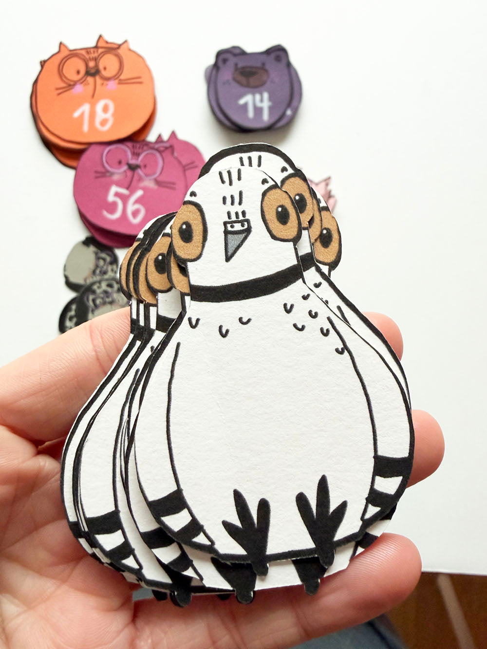 Hand hält Stapelspiel im Tauben-Design von Halfbird mit anderen Kartendecks, wie Katze und Bär im Hintergrund