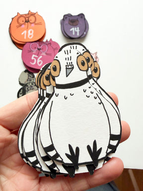Hand hält Stapelspiel im Tauben-Design von Halfbird mit anderen Kartendecks, wie Katze und Bär im Hintergrund