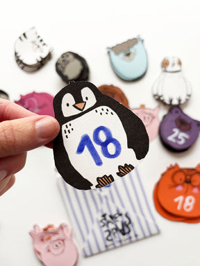 Hand hält Pinguin Spielkarte mit der Zahl 18 aus dem Spielkartendeck von Halfbird. Im Hintergrund sichtbar, der gestreifte Umschlag und andere Kartenstapel, wie Schwein, Katze, Bär, Lama, Taube, Katze,...