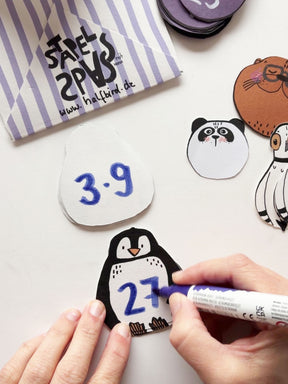 Hand malt mit einem blauen Acrylstift Rechenaufgaben und -ergebnisse auf die Stapelkarten von Halfbird im Pinguin-Design.