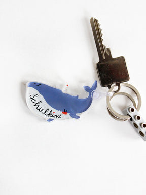 Ein Schlüsselanhänger in Form eines Wals mit Schriftzug Schulkind in den Farben blau und weiß mit Partyhut