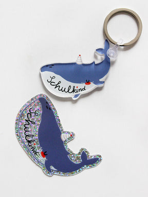 Ein Schlüsselanhänger und ein Glitzer Sticker in Form eines Wals mit Schriftzug Schulkind in den Farben blau und weiß mit Partyhut