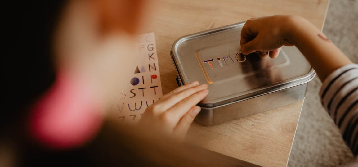 Kind klebt spülmaschinenfeste Namenssticker ABC auf eine Brotdose