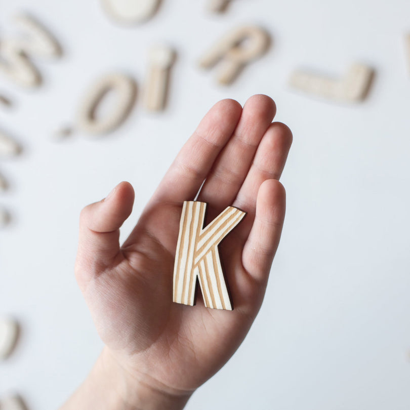 Kinderhand hält einen Holzbuchstaben K mit streifen, dahinter liegen noch mehr Holzbuchstaben