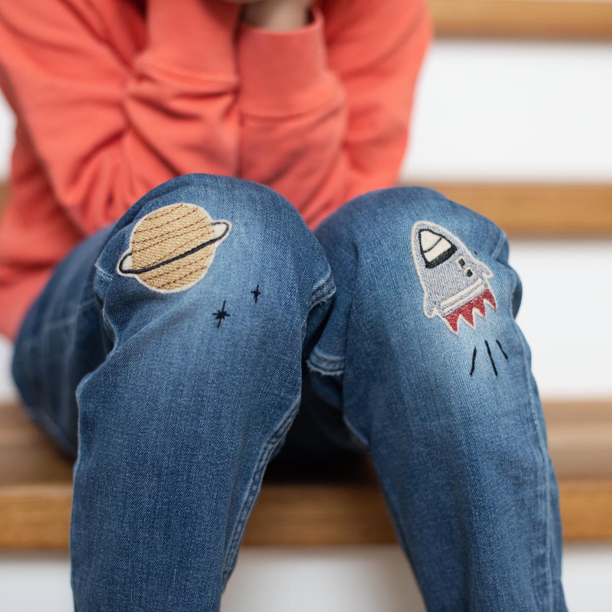 Kind sitzt auf der Treppe und hat eine geflickte Jeans Hose an. Auf den Knien sind zwei Bügelflicken von halfbird, ein Planet in gold und eine Rakete in grau