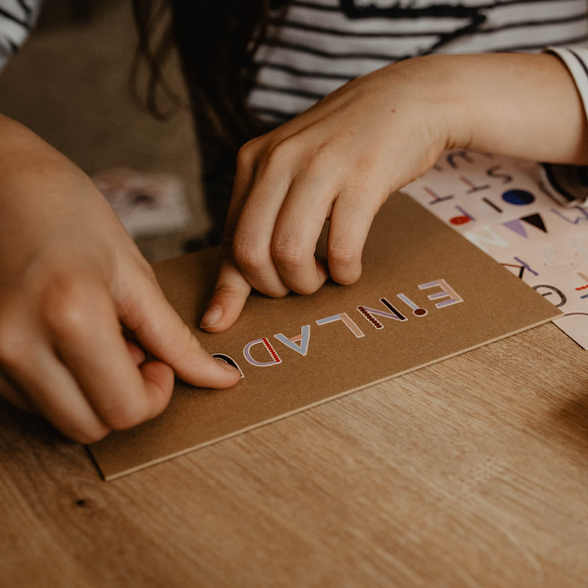 Kind klebt Buchstabensticker auf eine Klappkarte mit dem Wortlaut Einladung