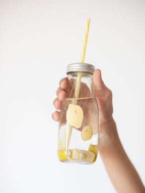 eine Kinderhand hält ein trinkglas mit gelbem strohhalm hoch, darauf kleben zwei zitronen als spülmaschinenfeste sticker und darin ist wasser