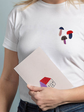 Frau mit weißem T-shirt und Pilz Aufnähern auf der Brust, sie hält ein Journal mit gesticktem Aufnäher Haus in der Hand