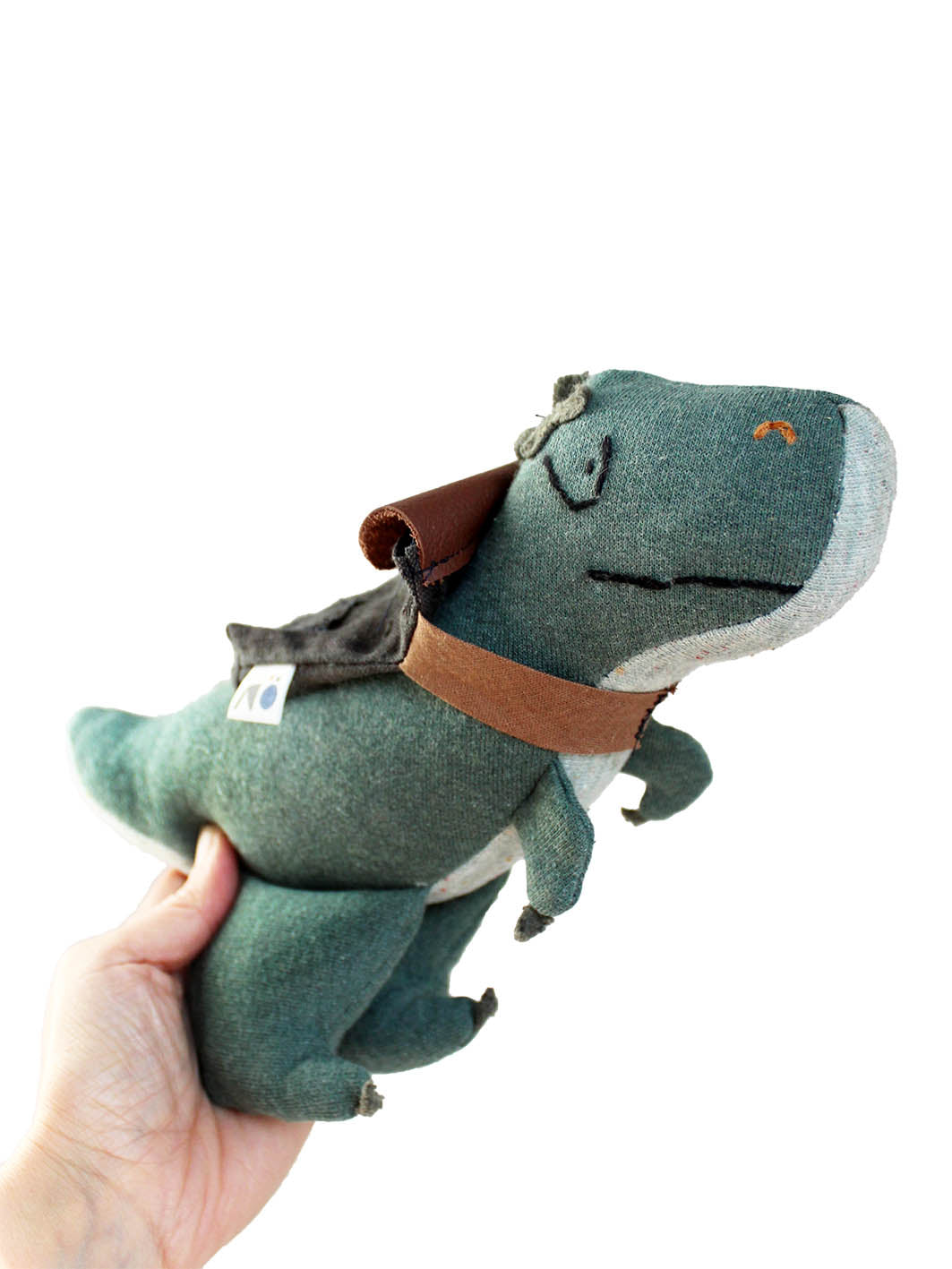 Hand hält ein Dino Kuscheltier mit einem Rucksack auf dem Rücken hoch. Der T-rex ist grün